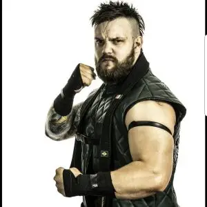 The man known as fowler wearing a green sleeveless jacket with black tape at the top of his biceps and taped hands with a black belt around his neck.