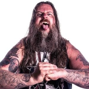 Dean is standing with is tongue out fists together in a sleeveless shirt and has many tattoos on his arms.