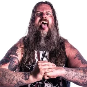 Dean is standing with is tongue out fists together in a sleeveless shirt and has many tattoos on his arms.