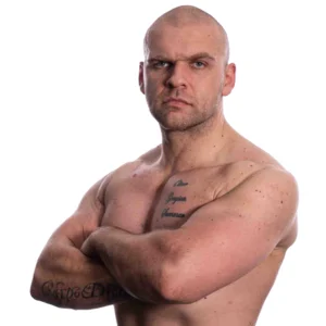 Cy stands shirtless with tattoos visible and his arms folded